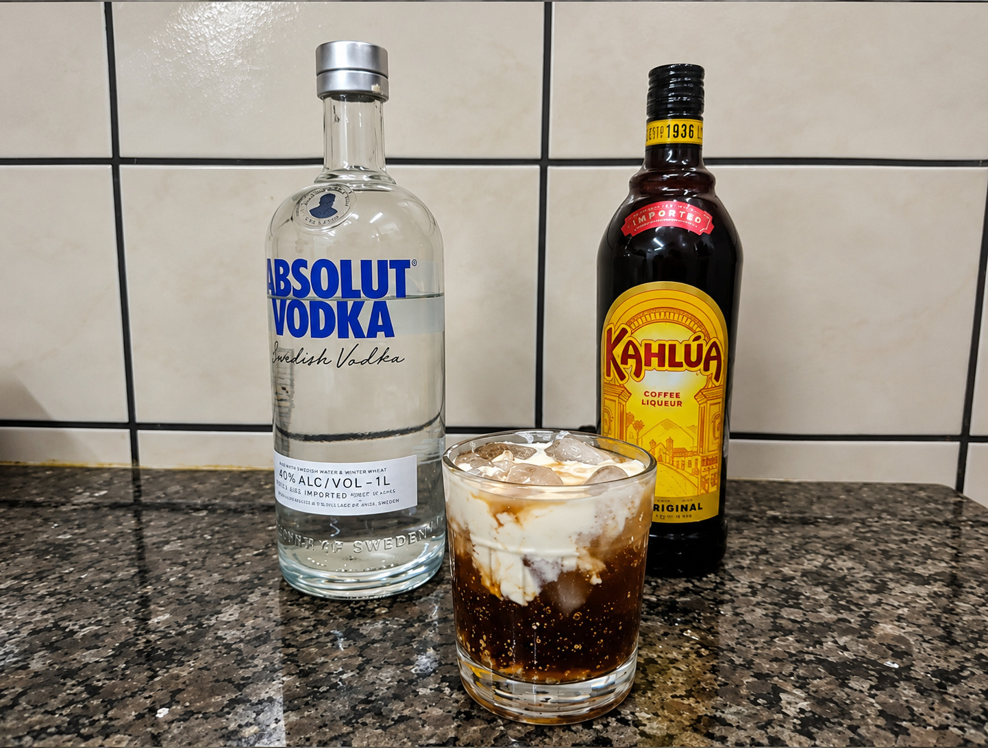White Russian com creme e gelo, com garrafas ao fundo.